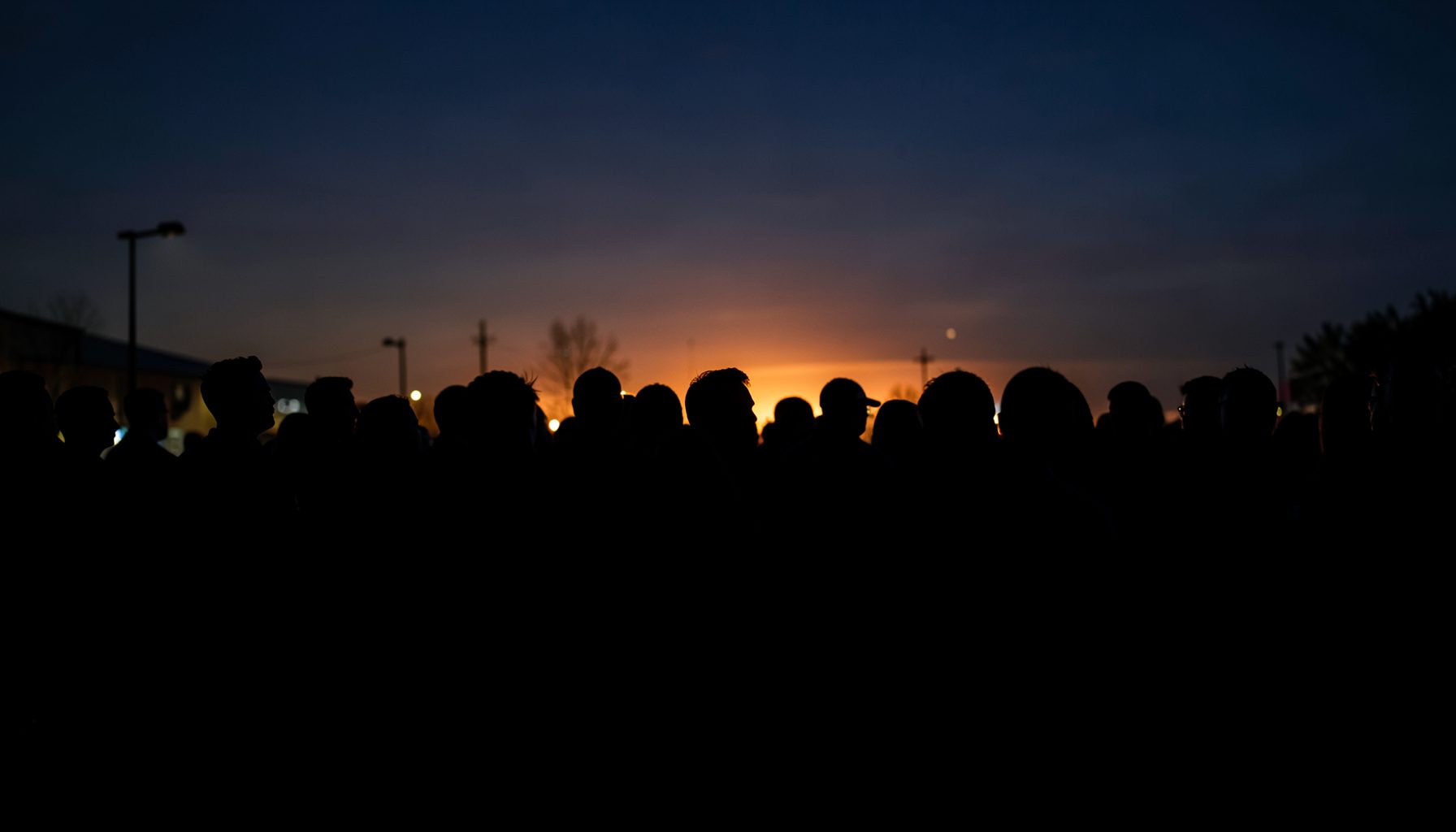Dramatic high-contrast silhouette of a crowd gathered at dusk, deep navy and black tones with warm amber light breaking through