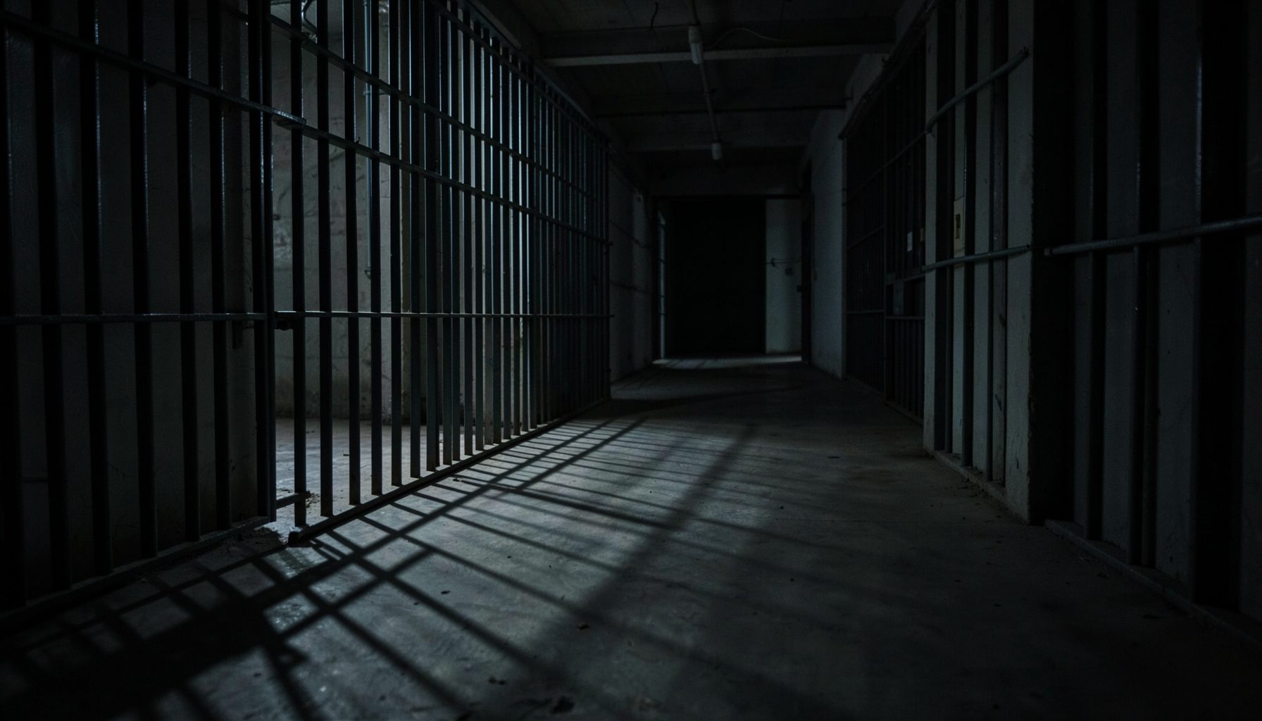 Dimly lit interior of a prison corridor with metal bars casting long shadows across a concrete floor