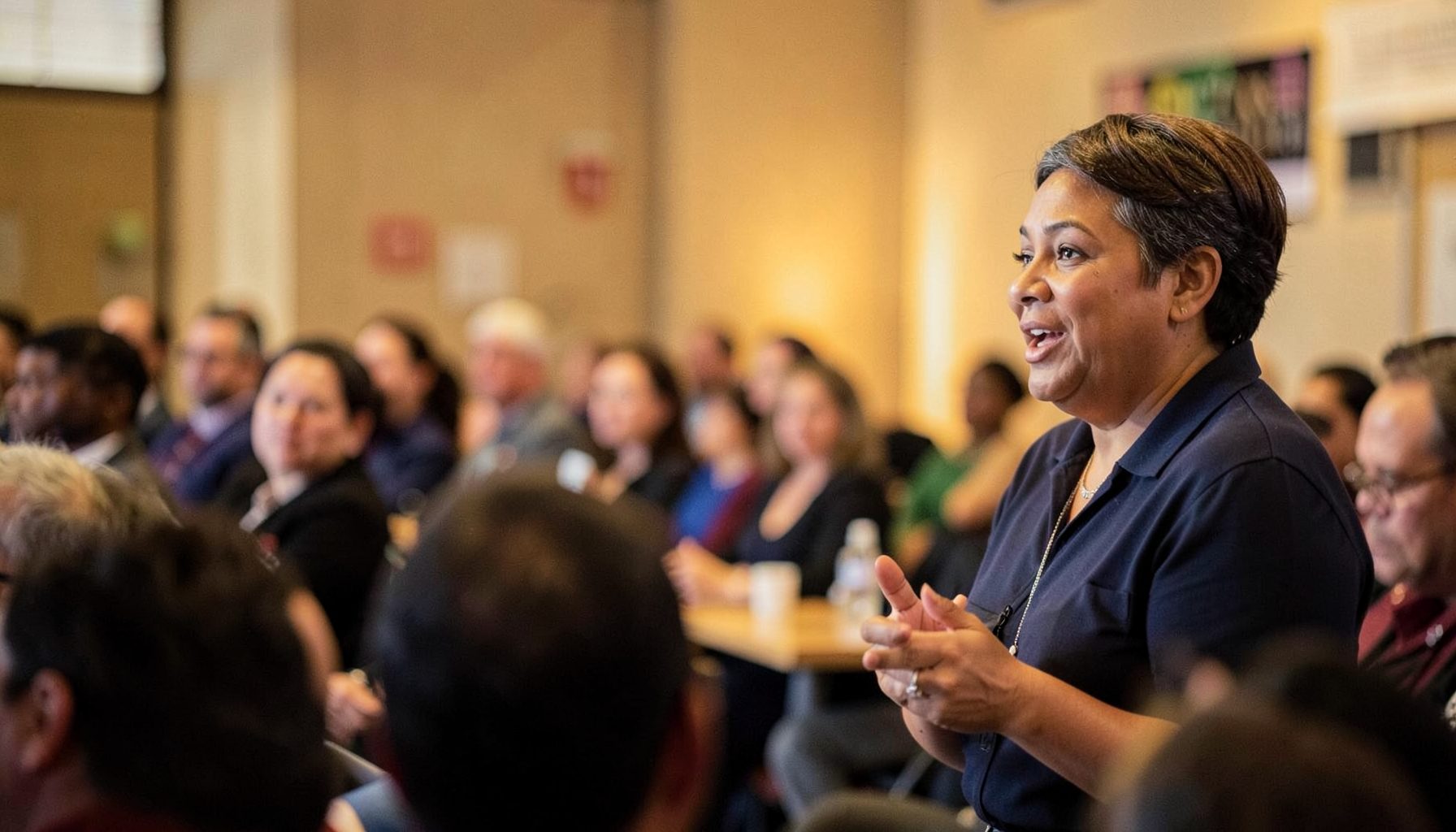 A person speaking at a community forum, warm indoor lighting, engaged audience in soft focus, advocacy and public dialogue