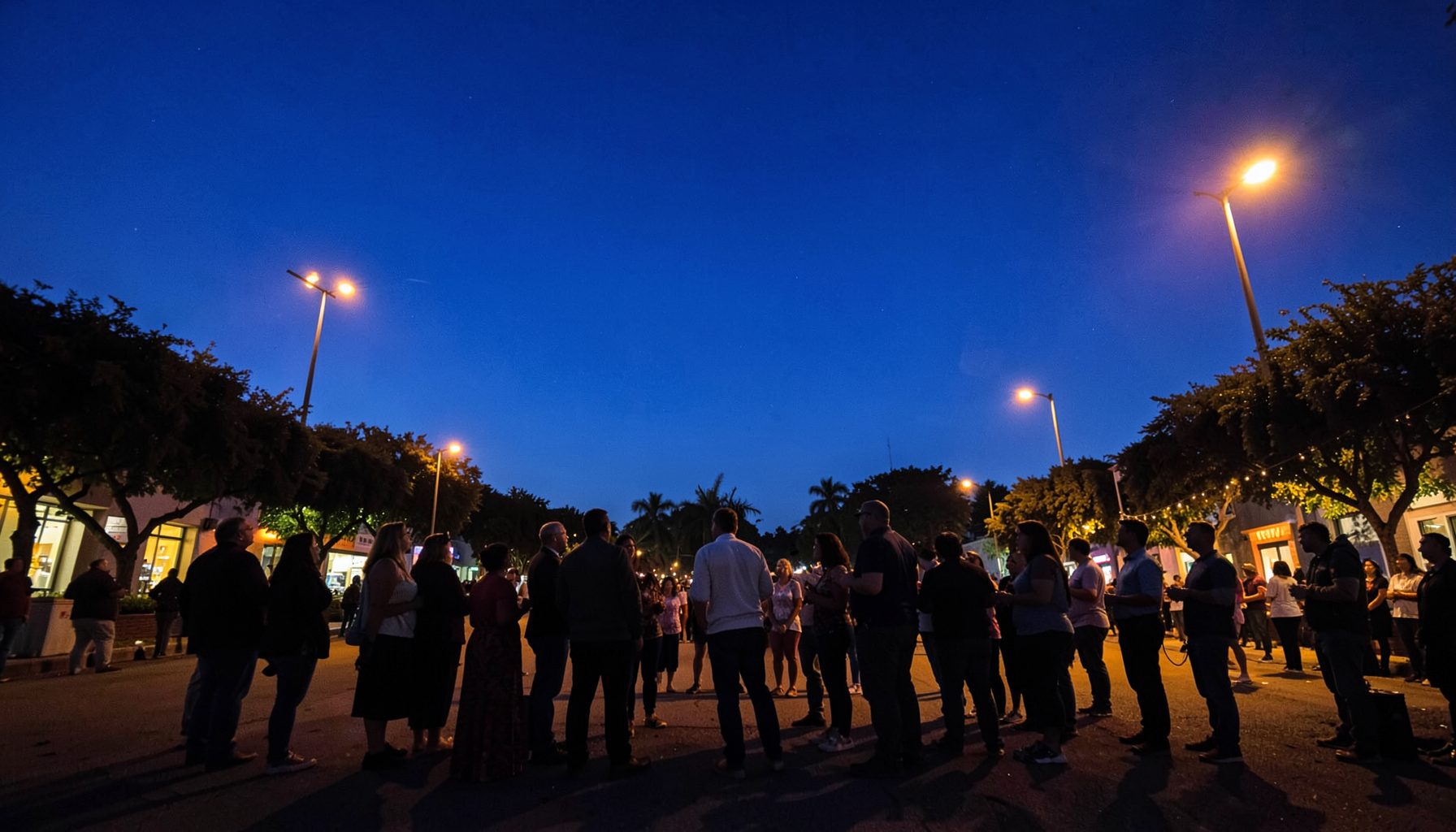 Wide-angle view of a peaceful community gathering at twilight, deep blue sky with warm streetlights, silhouetted figures standing together